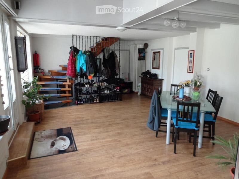Open-plan living and dining area with wooden flooring, a glass dining table with blue chairs, a staircase leading to an upper level, and a coat rack with jackets and shoes.