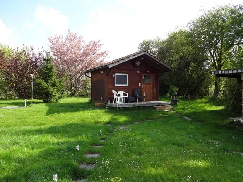 A small wooden cabin with a pitched roof and a single window sits on a grassy lawn surrounded by trees. A small wooden deck with two chairs and a table is attached to the front of the cabin.