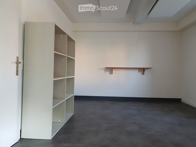 Empty room with a grey carpeted floor, white walls, a white shelving unit, and a small wooden shelf on the wall.