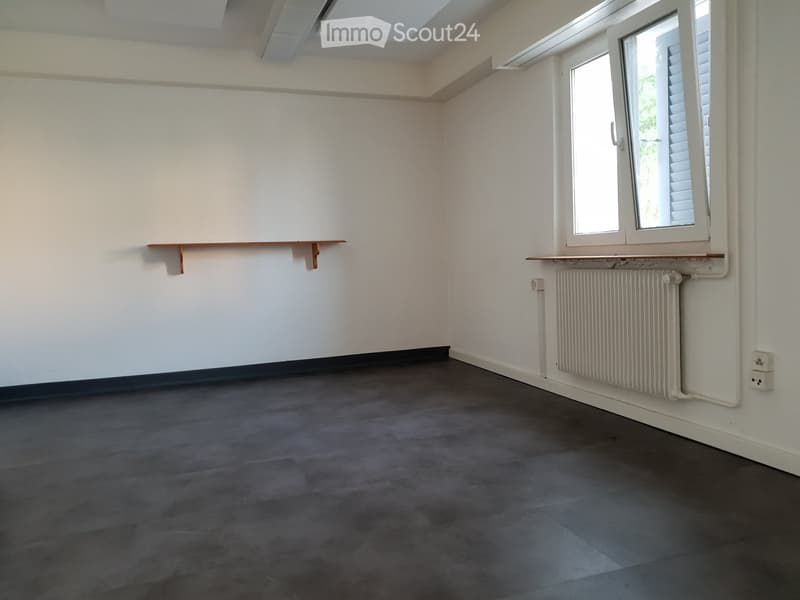 Empty room with white walls, dark grey carpeted floor, a window with blinds, a radiator, and a small wooden shelf attached to the wall.