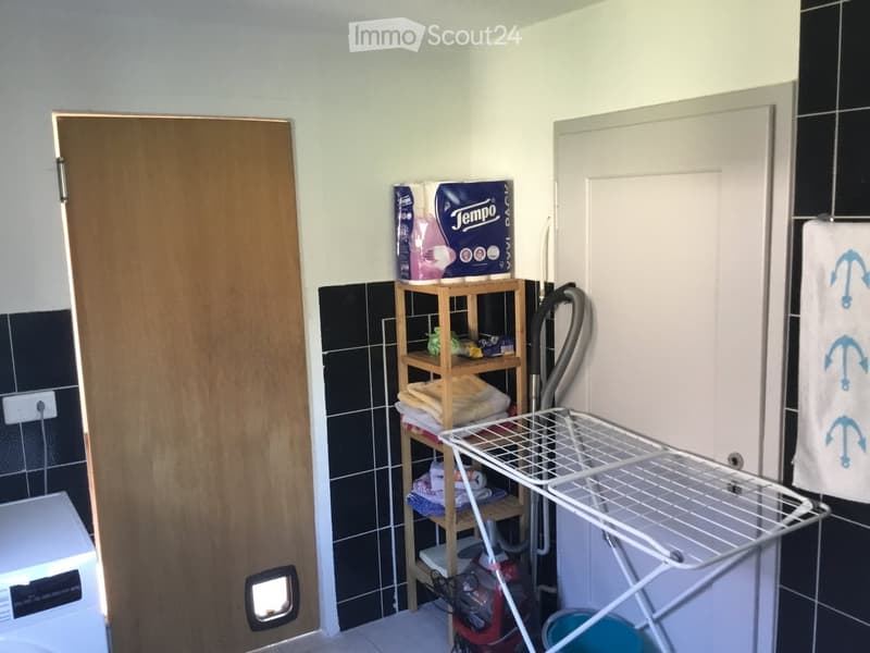 Bathroom with a wooden shelf holding towels and laundry supplies, a white laundry rack, a washing machine, and a vacuum cleaner in the corner.