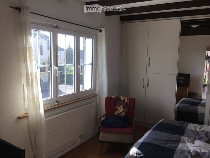 Bright bedroom with wooden flooring, large window with white curtains, a red armchair with a blanket, a bed with a patterned quilt, and a white wardrobe with mirrored doors.
