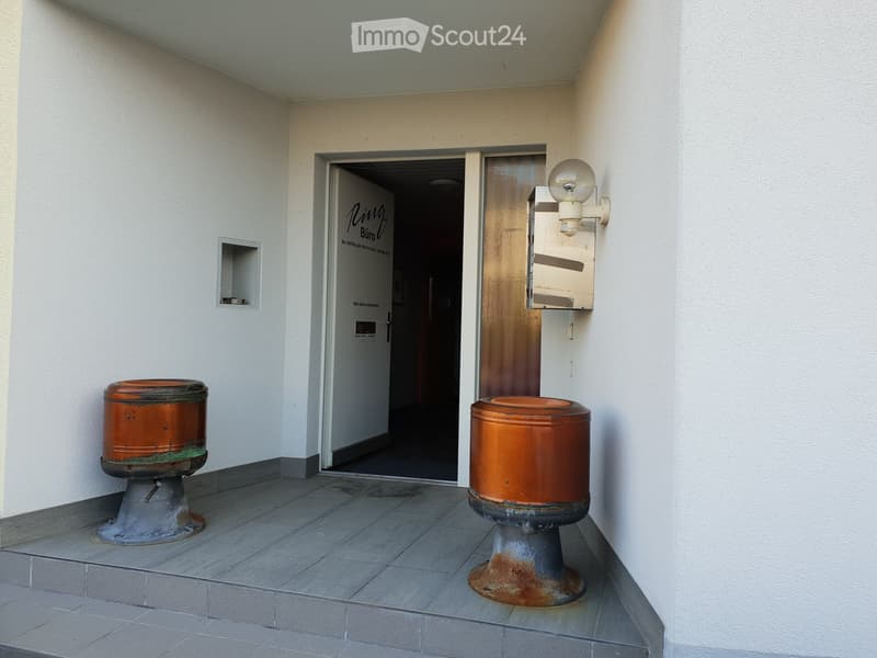 A hallway with white walls, a tiled floor, and two orange cylindrical objects on stands. A door leads to another room labeled "Ring Büro."