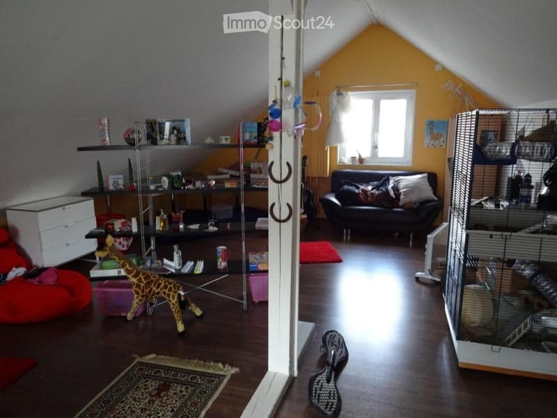 Attic room with slanted yellow walls, wooden floor, large window, black sofa, shelves with books and decor, small cage, toy giraffe, bean bag chair, and area rug.
