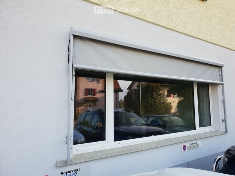 Window with a partially retracted roller shade, showing reflections of parked cars and neighboring buildings.