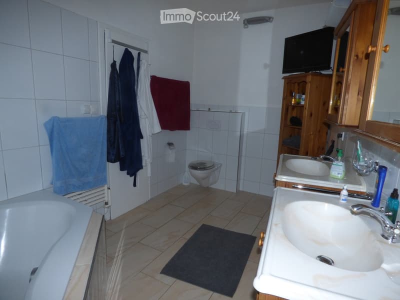Bathroom with a bathtub, toilet, sink, mirror, and wooden cabinet. Towels are hanging on the wall, and a small TV is mounted above the cabinet.