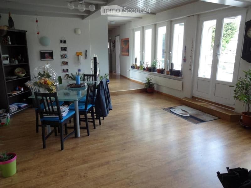 Bright, open-plan living and dining area with wooden flooring, large windows, a dining table with chairs, a bookshelf, and potted plants.