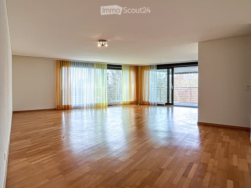 Empty room with wooden flooring, large windows with curtains, and a sliding glass door leading to a balcony.