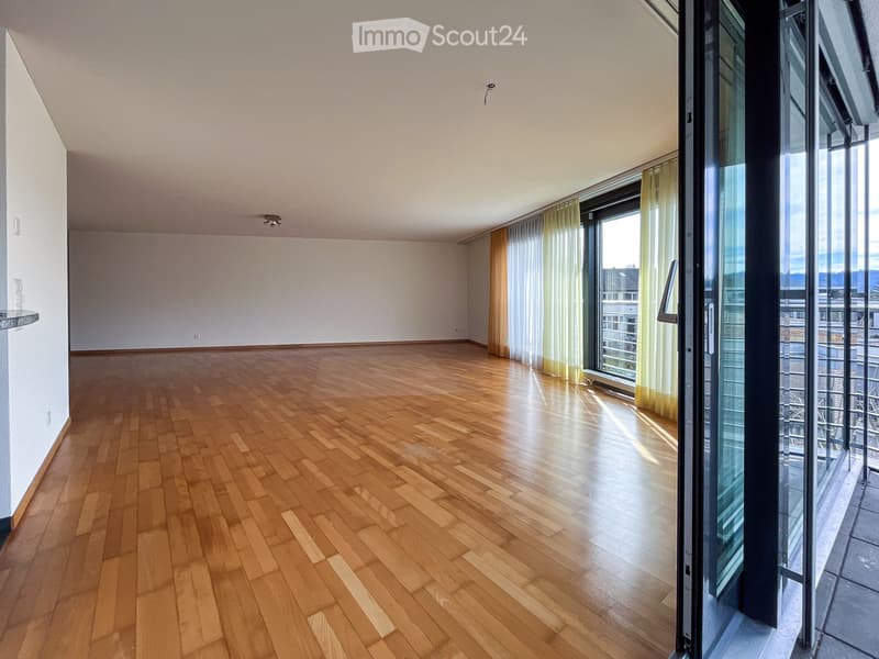Empty room with wooden flooring, white walls, large windows with sheer curtains, and a sliding glass door leading to a balcony.