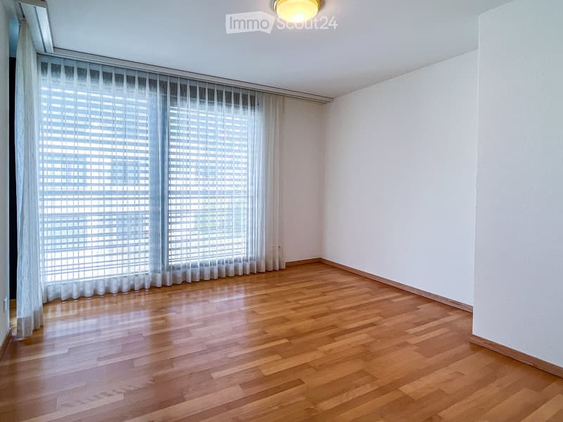 Empty room with wooden flooring, white walls, large window with white blinds, and a ceiling light fixture.
