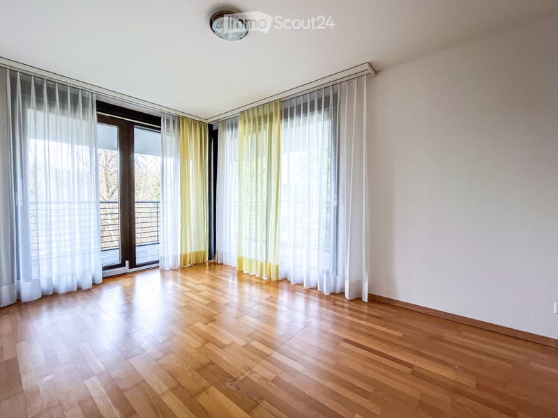 Empty room with wooden flooring, large windows with white and yellow curtains, and a balcony visible through the windows.