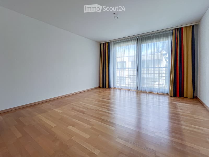 Empty room with wooden flooring and white walls, featuring large windows with sheer curtains and colorful drapes.