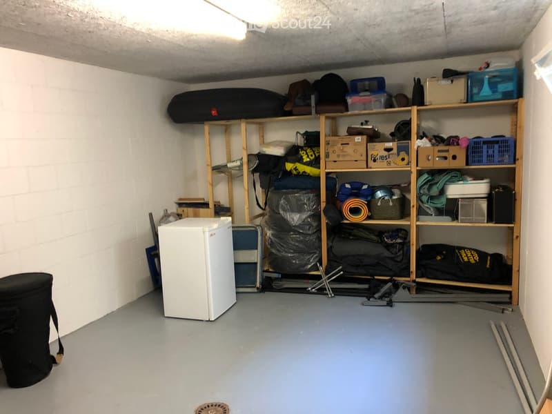Storage room with wooden shelves filled with bags, boxes, and exercise equipment, a small refrigerator, and a black trash bin.