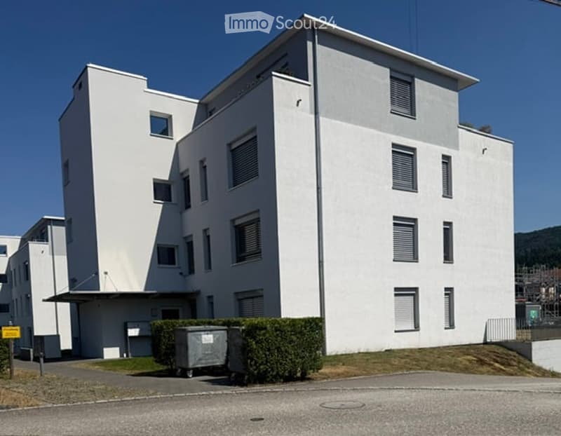 Modern, multi-story white building with rectangular windows and a flat roof, surrounded by a small landscaped area with hedges and a trash bin.