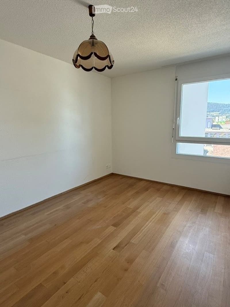 Empty room with wooden flooring, white walls, and a single window with blinds. A hanging light fixture is visible from the ceiling.