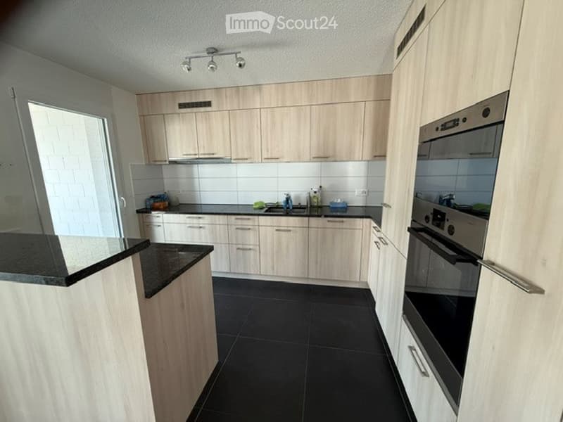 Modern kitchen with light wooden cabinets, black countertops, and black tiled flooring. Features a sink, oven, and various small appliances.
