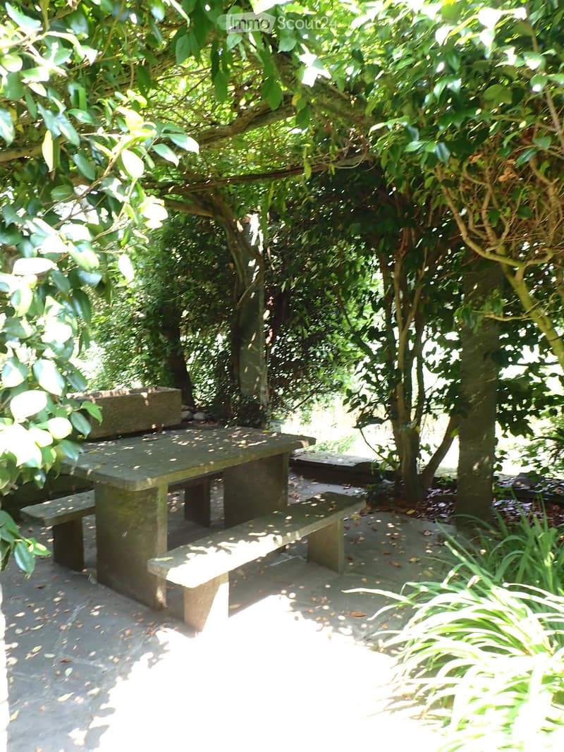 A serene garden area with stone benches, lush greenery, and trees providing shade.
