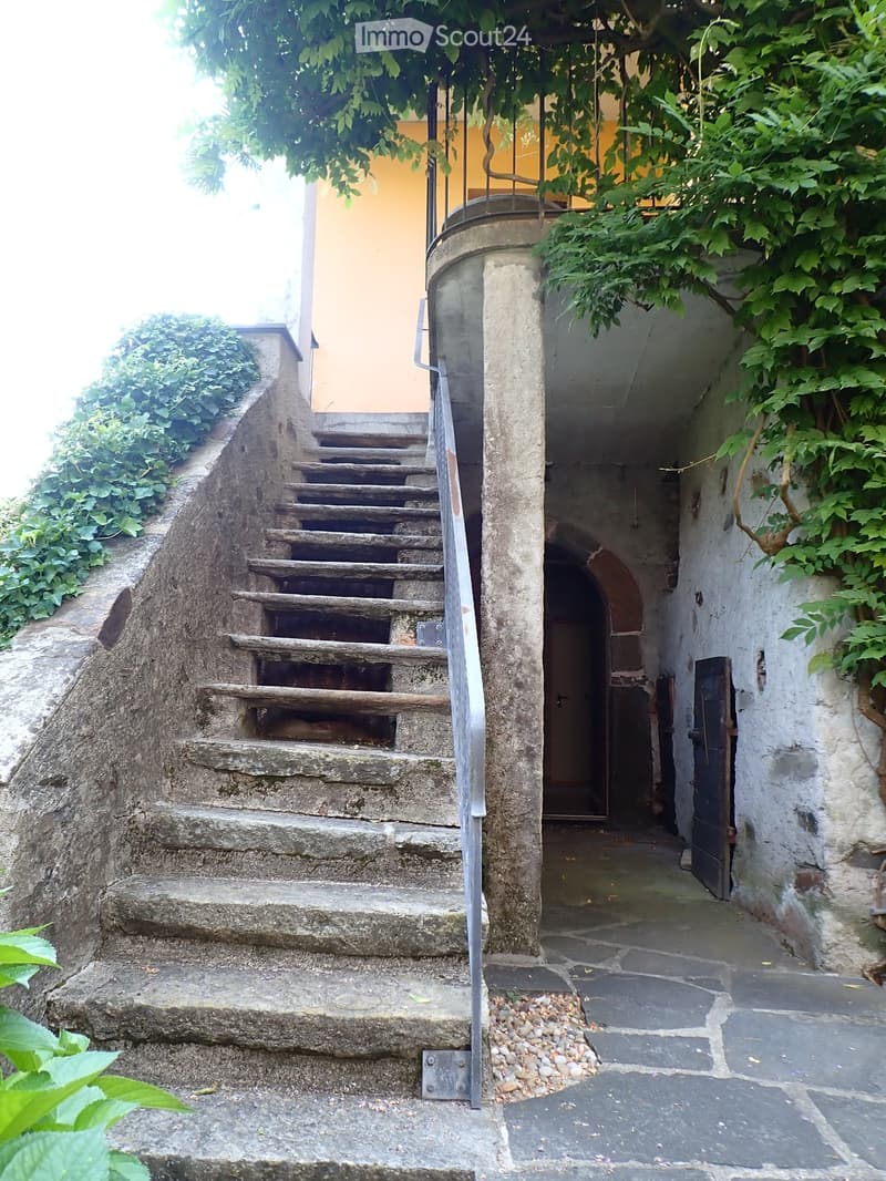 A narrow stone staircase leads up to a doorway, flanked by stone walls and greenery, with a metal railing on one side.
