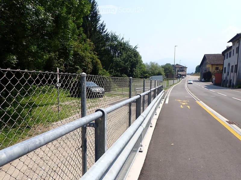 A paved pathway bordered by a metal fence, with a road visible in the background and trees lining the area.