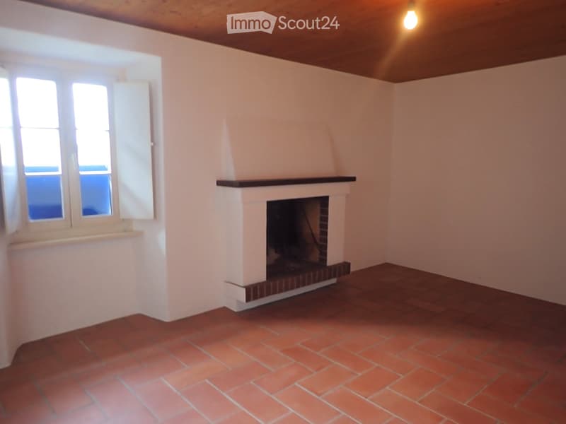 Empty room with red tiled floor, white walls, and a fireplace with a wooden mantel. The room has large windows with shutters and a wooden ceiling.