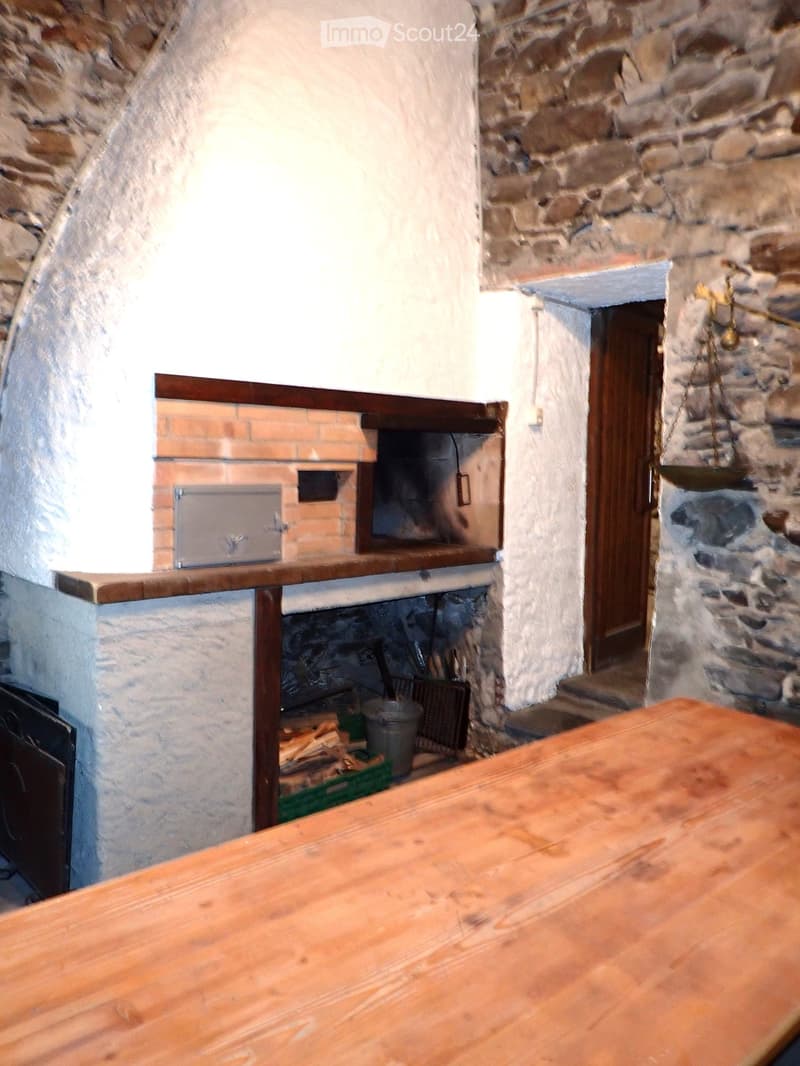 A rustic room with a large wooden table, a stone fireplace with a wooden shelf above it, and a wooden door leading to another room.