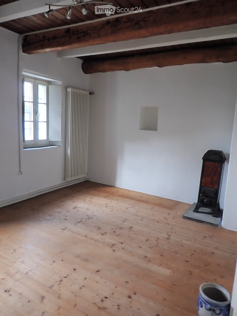 Empty room with wooden flooring, white walls, exposed wooden beams on the ceiling, a radiator, and a window with shutters.