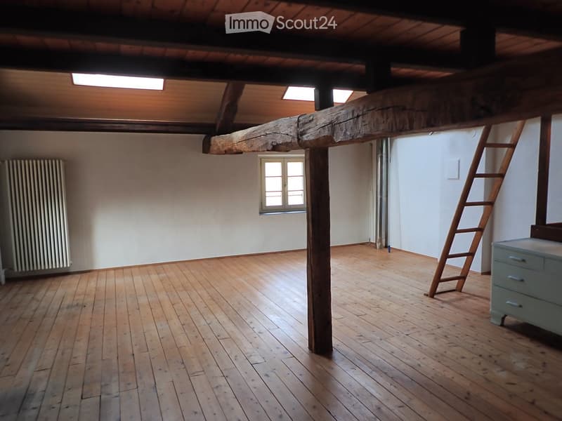Empty room with wooden floor, exposed wooden beam, window, radiator, ladder, and small cabinet.