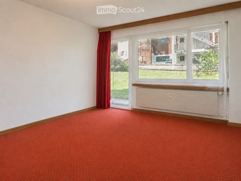 Empty room with red carpet, white walls, wooden trim, large windows with curtains, and a radiator below the windows.