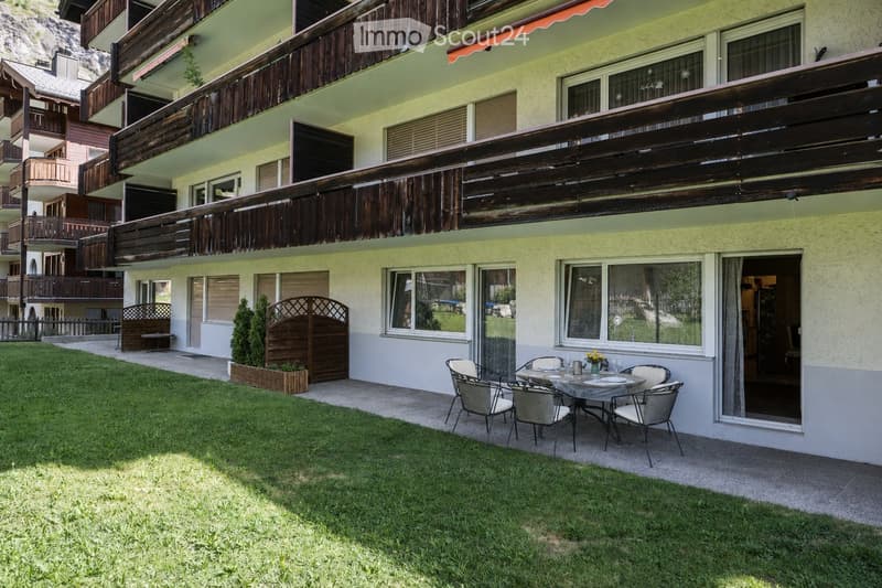 Garden area with a small table and chairs, surrounded by a grassy lawn and a building with balconies and large windows.
