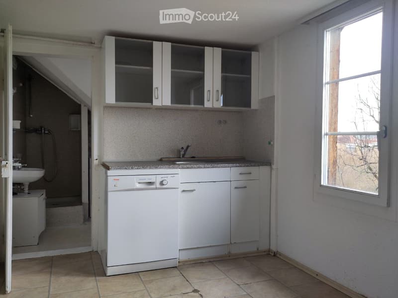 A small, modern kitchen with white cabinets, a dishwasher, a sink, and a mirror above the sink. The room has tiled flooring and a window letting in natural light.
