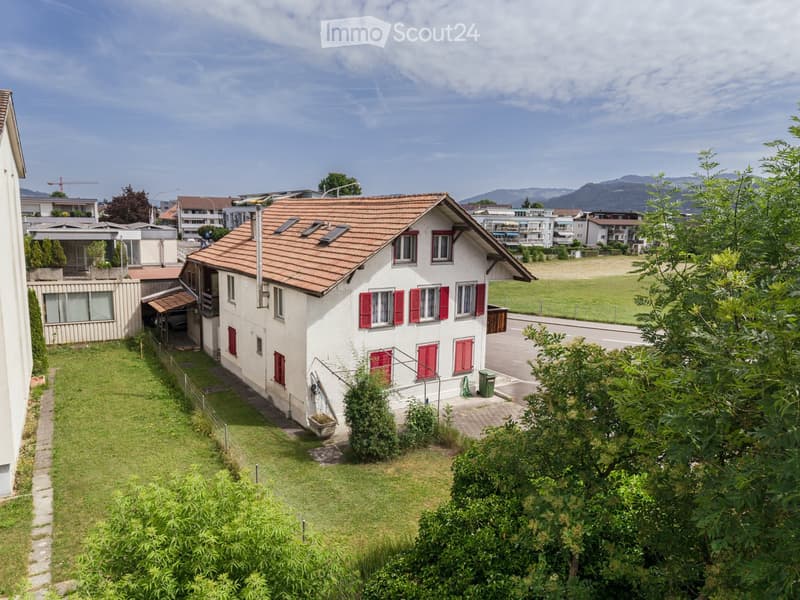 A two-story house with a red-tiled roof and white walls, featuring red shutters on the windows. The house is surrounded by a well-maintained lawn and trees, with a paved driveway and a small garden area.