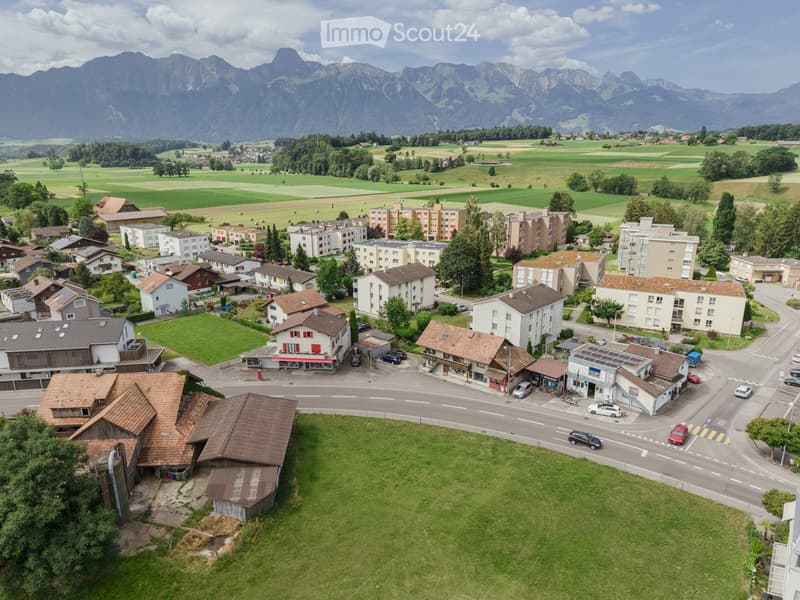 Aerial view of a small town with a mix of residential and commercial buildings, surrounded by green fields and mountains in the background.