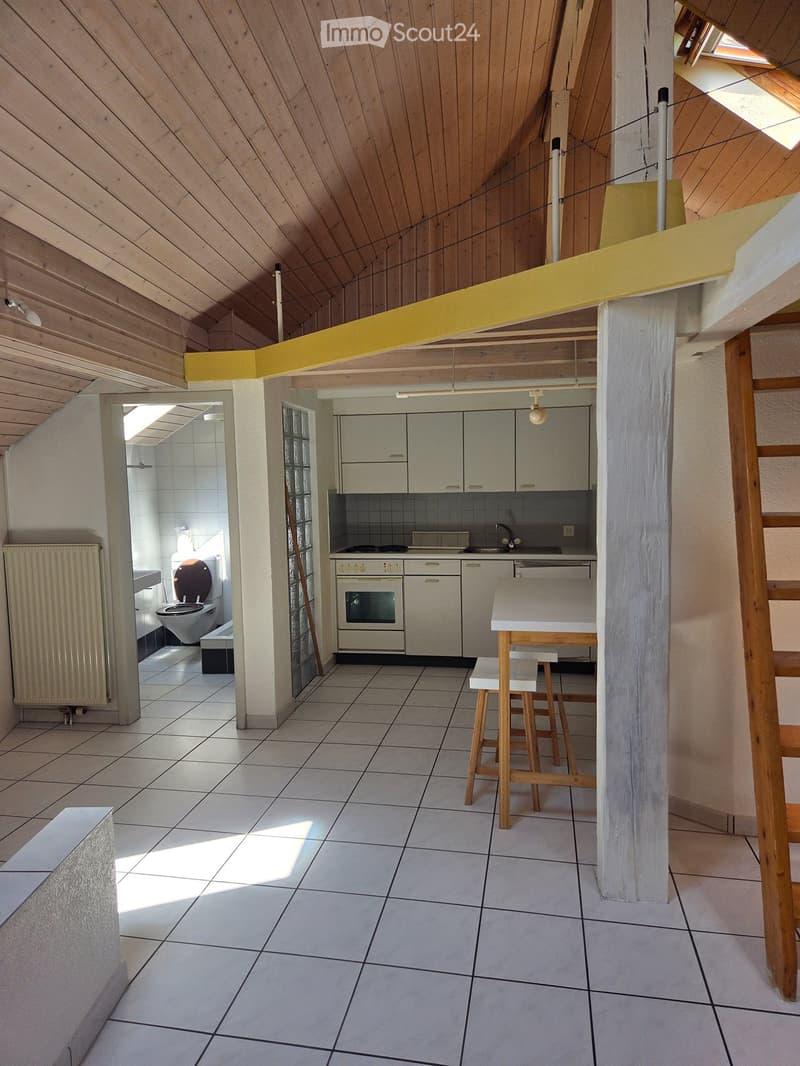 Open-plan space with a kitchen area, bathroom, and living area. Kitchen features white cabinets, a sink, and an oven. Bathroom includes a toilet and radiator. Wooden stools are placed near the kitchen counter.