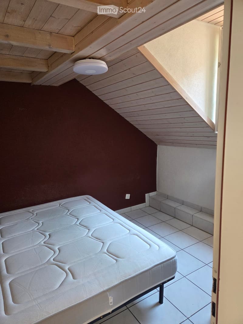 Empty bedroom with a white mattress on a bed frame, wooden slanted ceiling, tiled floor, and a partially open door leading to another room.