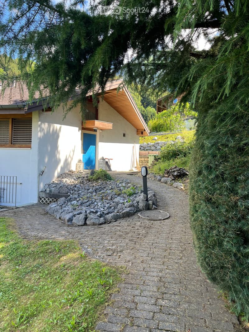 A modern two-story house with a light-colored exterior, a blue front door, and a stone pathway leading to the entrance, surrounded by greenery and trees.