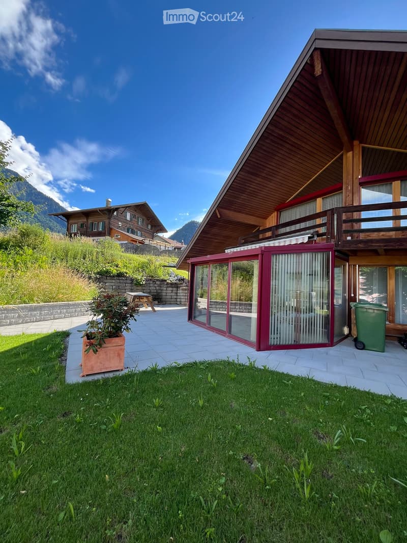 A modern wooden house with a sloped roof, large windows, and a balcony overlooking a grassy lawn and a stone wall. The house is surrounded by a well-maintained garden and a scenic mountain backdrop.