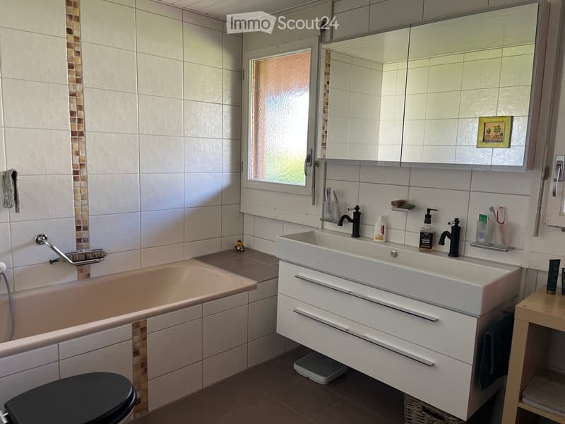 Modern bathroom with a bathtub, double sink, large mirror, and a toilet. The walls are tiled with a decorative mosaic border, and the floor is tiled in a warm brown color. A small window provides natural light.