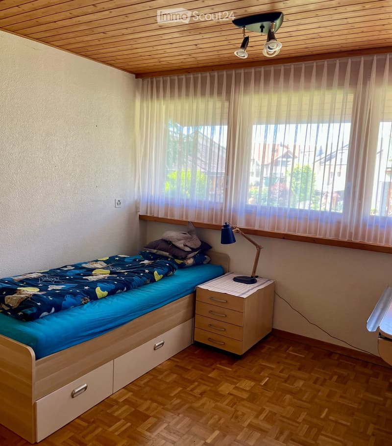 Cozy bedroom with a single bed, nightstand, and lamp. The bed has a blue duvet and pillows, and the room features wooden flooring and large windows with sheer curtains.