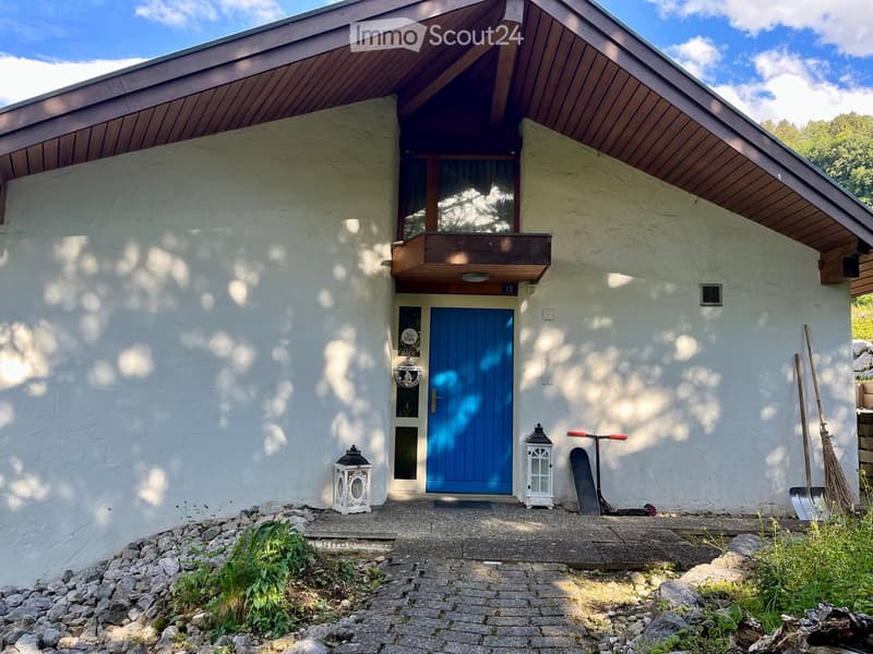 Single-story house with a blue front door, white walls, and a small balcony above the entrance. The house features decorative lanterns on either side of the door and a small garden area with rocks and plants.
