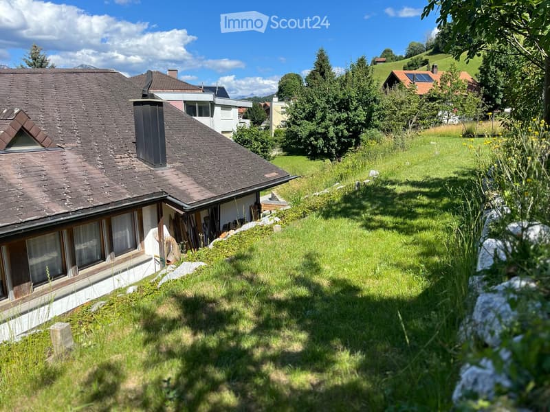 A two-story house with a dark shingled roof, white walls, and multiple windows, situated on a grassy slope with surrounding trees and hills in the background.