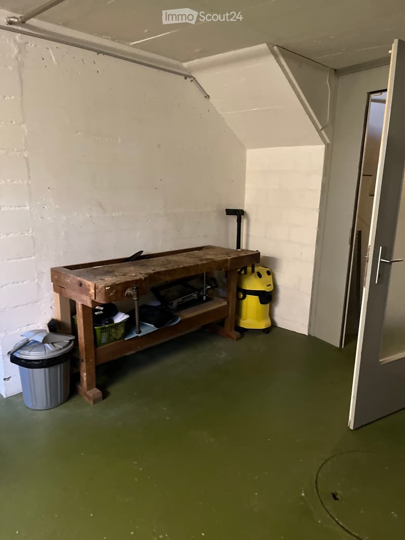 A small, utilitarian room with a workbench, vacuum cleaner, and trash can. The walls are white brick, and the floor is green.
