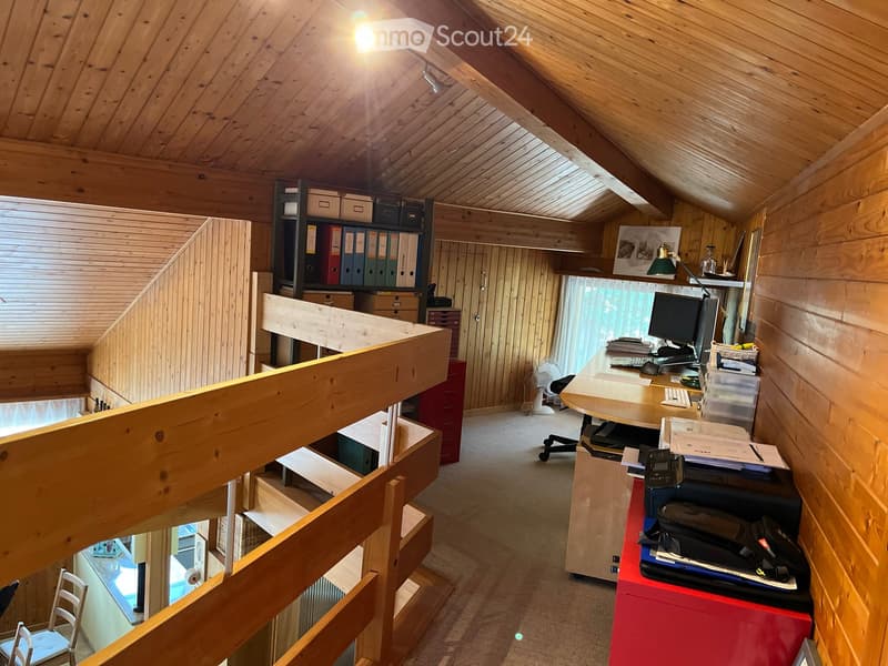 A well-lit attic office with wooden walls and ceiling, featuring a large desk with a computer, printer, and office supplies, a chair, a fan, and storage cabinets.