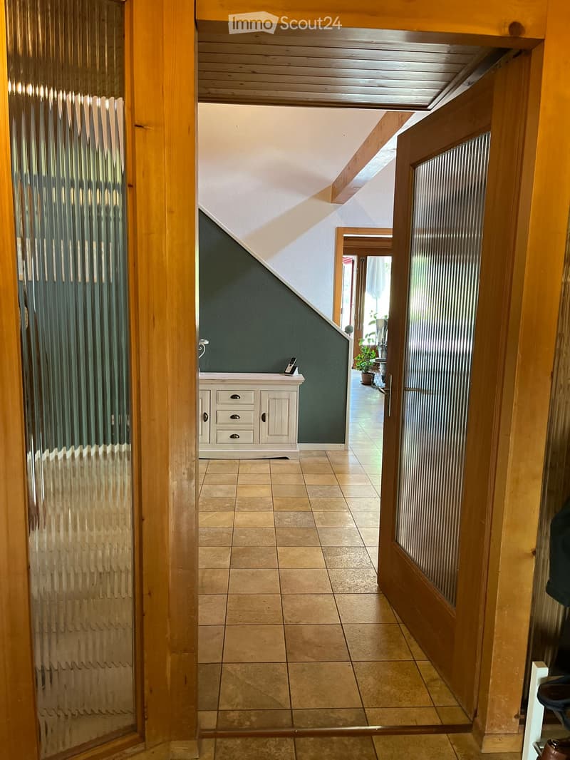 Hallway with tiled floor, wooden doors, and a small white cabinet with drawers and a phone on top. The hallway leads to another room with a visible plant and a partially open door.