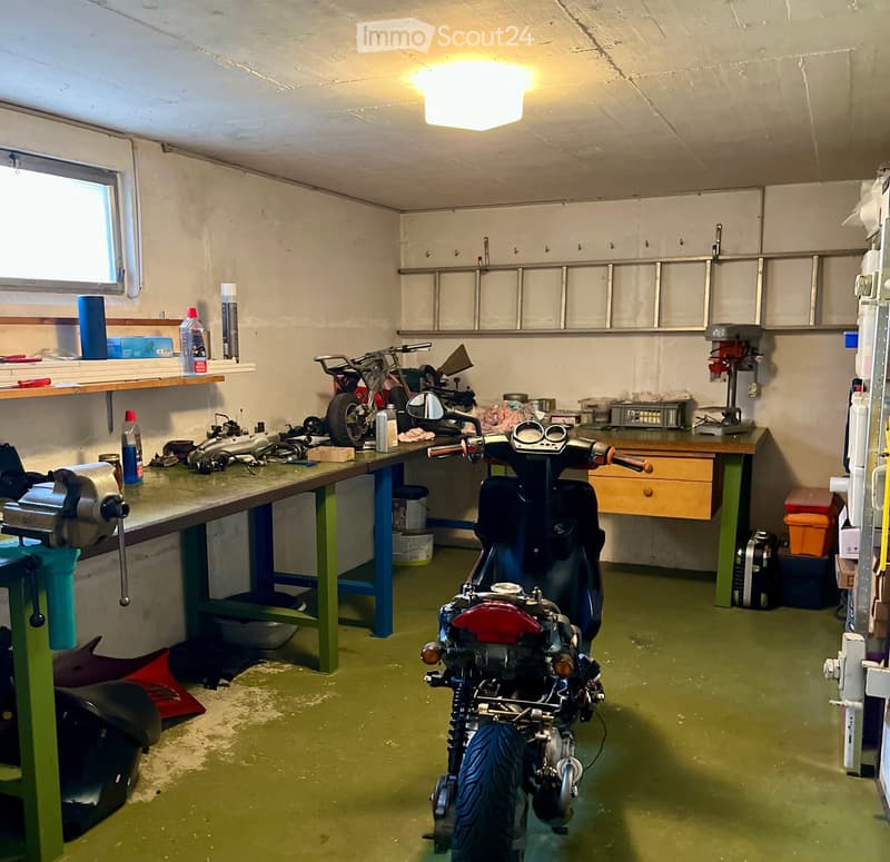 Garage with green flooring, white walls, and a motorcycle on a workbench. Shelves hold tools and supplies, and a drill press is visible.