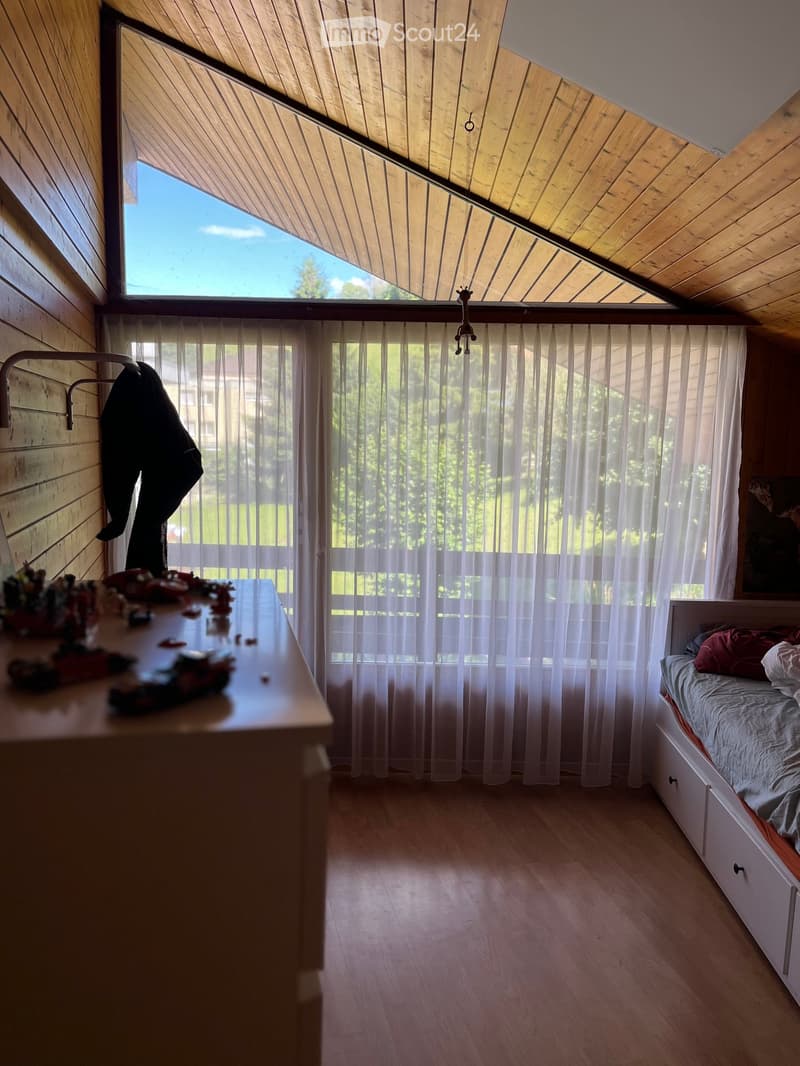 Cozy bedroom with wooden walls and ceiling, large window with sheer curtains, bed with pillows and blanket, nightstand, and small table with toys.