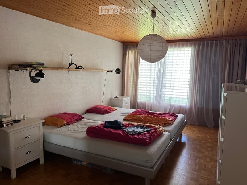 Cozy bedroom with wooden floors, slanted wooden ceiling, large window with curtains, double bed with colorful blankets and pillows, bedside tables with lamps, and a hanging paper lantern light fixture.