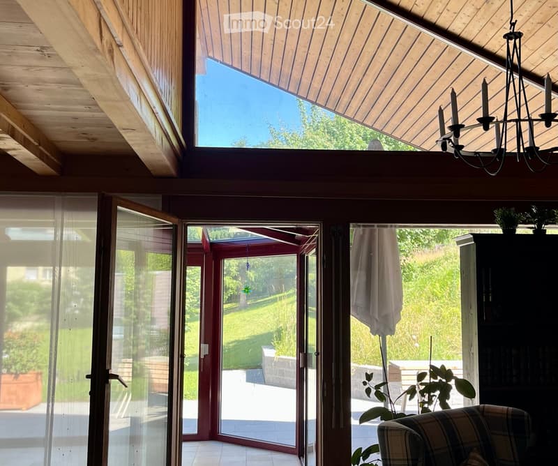 Bright living room with wooden ceiling, large skylight, and sliding glass doors leading to a garden. Features a patterned sofa, chandelier, and potted plants.