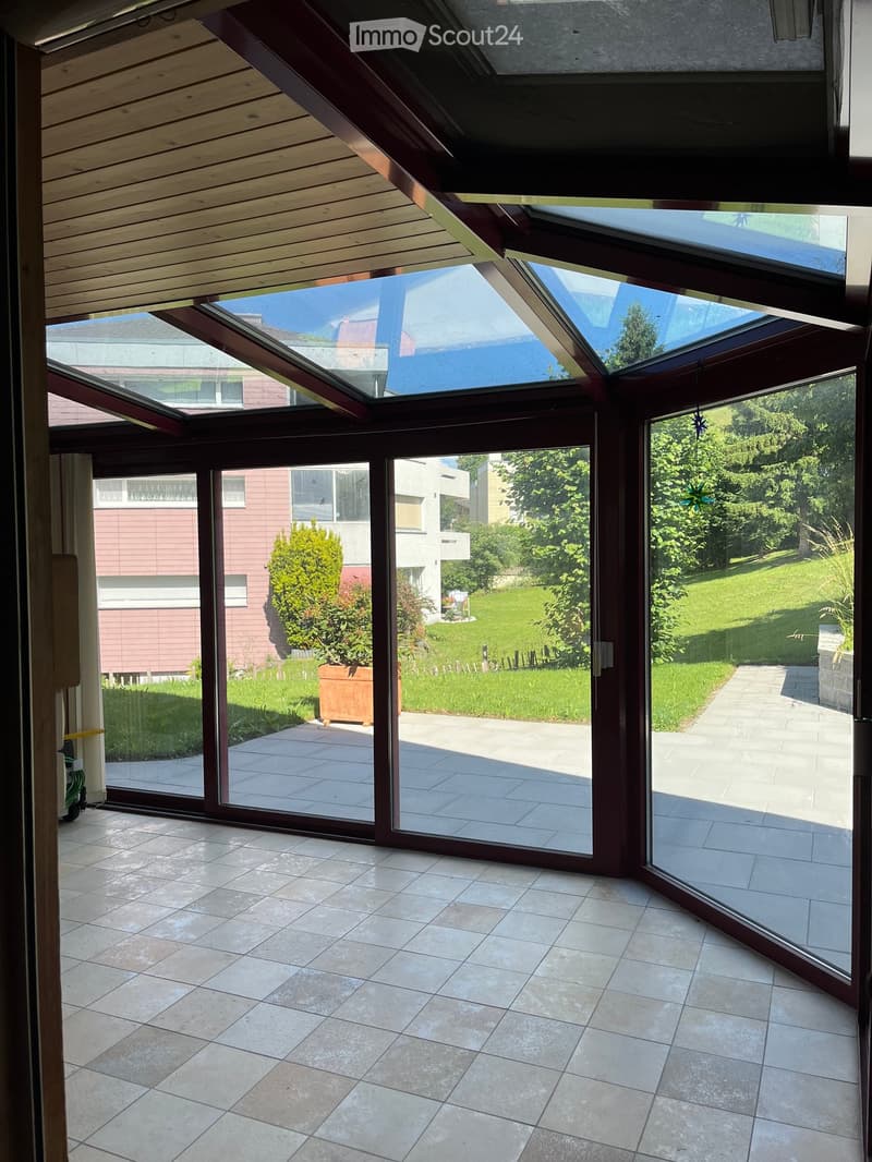 Bright, spacious sunroom with large glass sliding doors opening to a tiled patio and lush garden. The room features a wooden slanted ceiling and ample natural light.