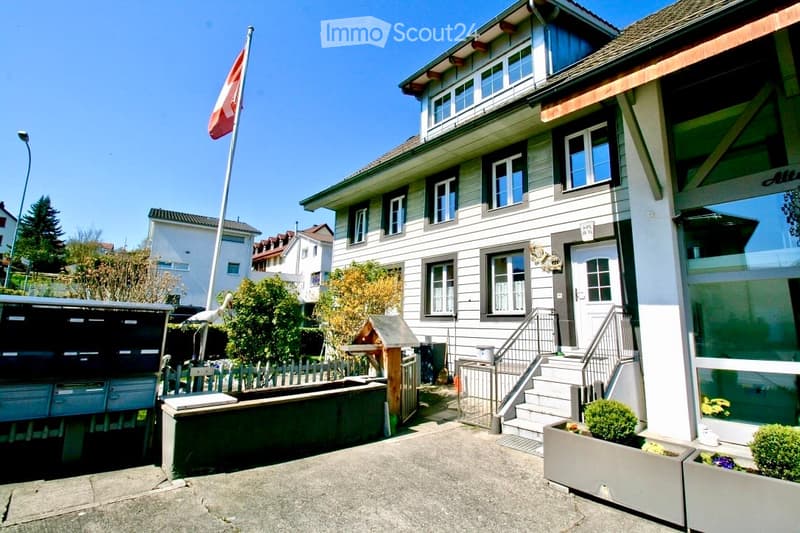 Two-story house with white siding, multiple windows, a front staircase, and a small garden area with potted plants.