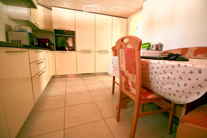 A modern kitchen with white cabinets, a tiled floor, and a small dining area featuring a wooden chair and a table with a floral tablecloth.
